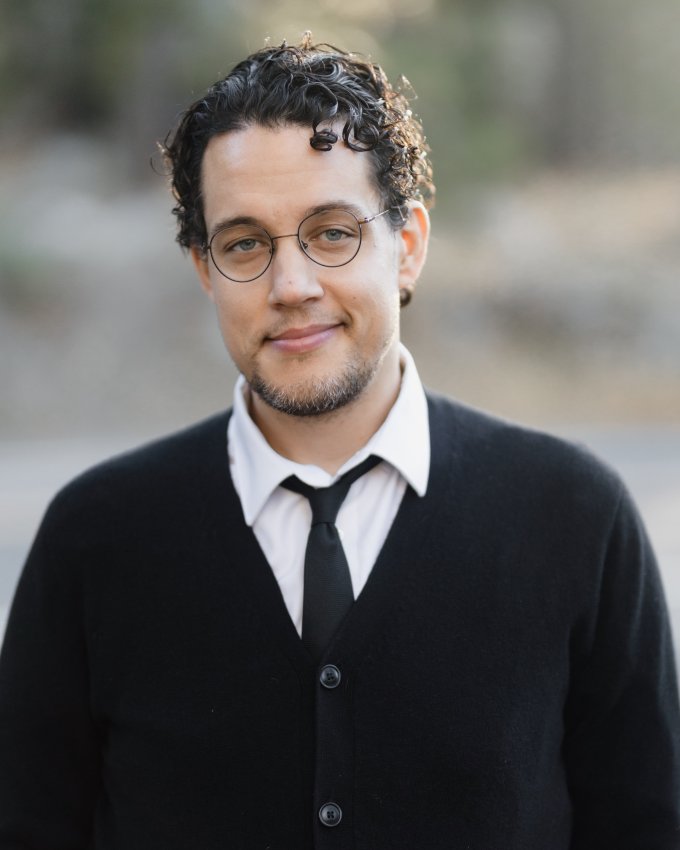 Portrait of a man with curly dark hair and glasses, wearing a black cardigan and tie, smiling softly outdoors