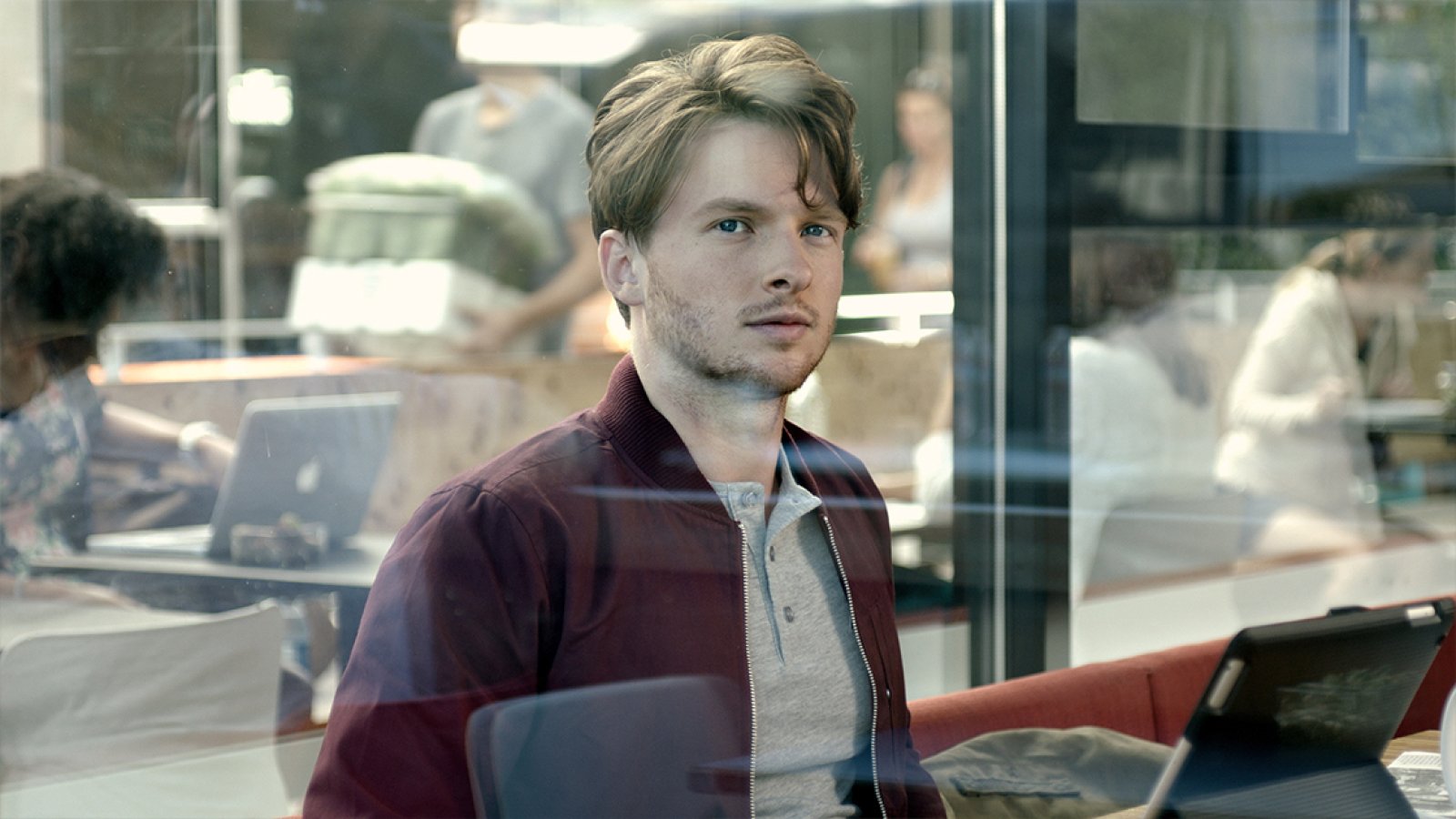 Young man in a maroon jacket seen through a cafe window, reflections of other patrons around him