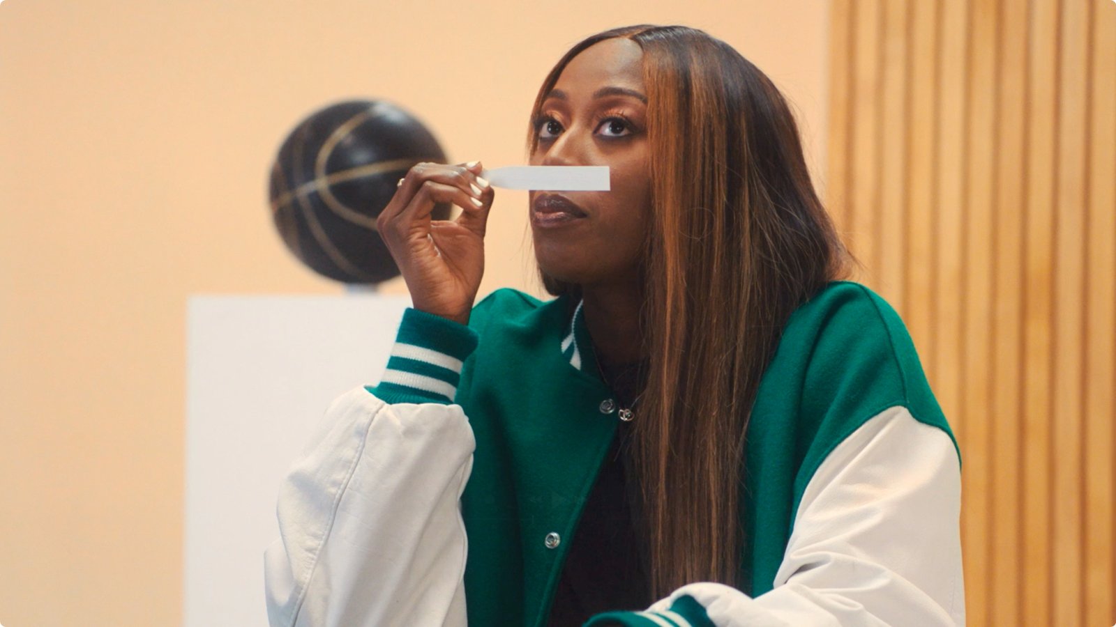 Woman in a green and white varsity jacket holding a nasal strip to her nose, basketball on a stand behind her