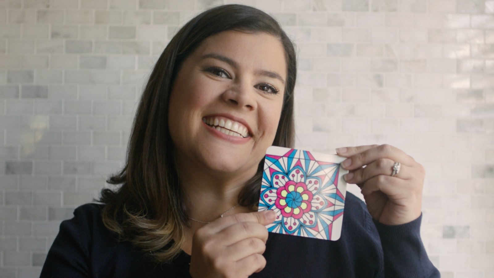 Smiling woman holding up a hand-colored mandala card against a white tiled wall