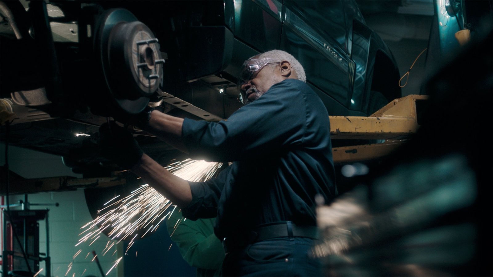 A mechanic in safety goggles grinds under a lifted truck as sparks fly across a dark garage