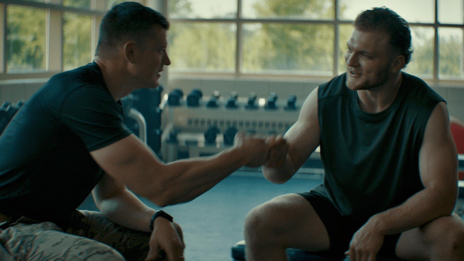 Two soldiers in workout gear fist-bumping in a gym, sunlit windows behind them