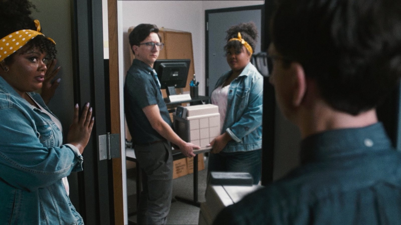 Coworkers in an office doorway carrying a boxy white 3D printer between them, others looking on