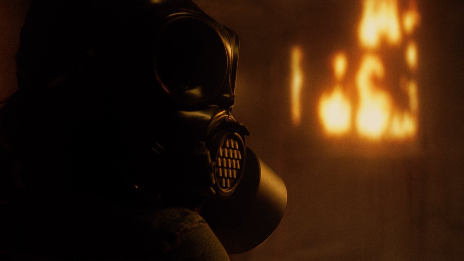 Silhouette of a figure in a gas mask watching flames burn in a dim, smoke-filled room