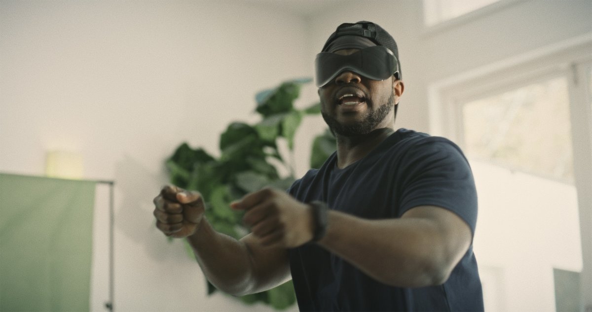 Man wearing a VR-style eye mask and cap, throwing punches in a bright room with a green plant behind him