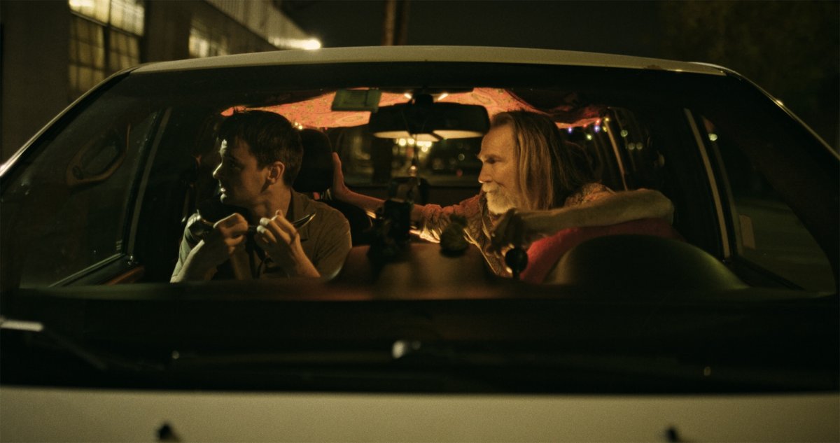 Two men seated in a parked car at night, one holding a small tool, string lights above them