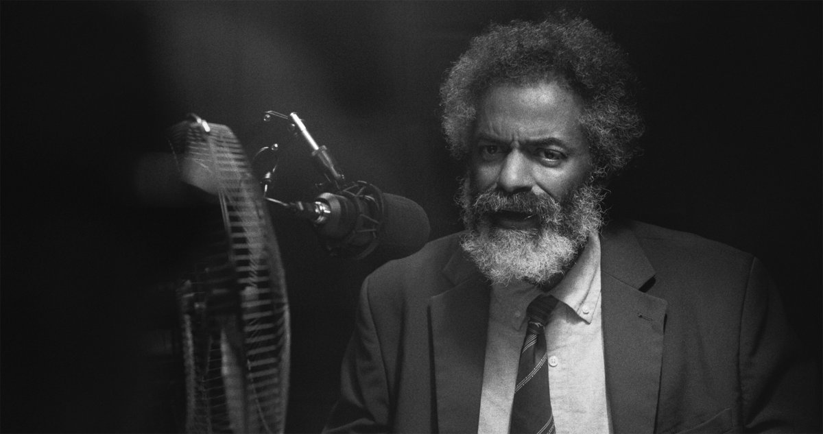 Black and white portrait of a bearded man in a suit and tie speaking into a podcast microphone