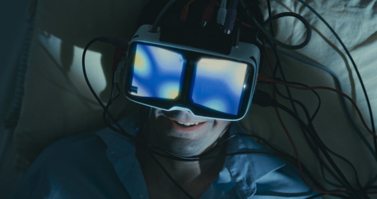 Smiling man lying down wearing a wired VR headset with glowing blue lenses, tangled cables around him