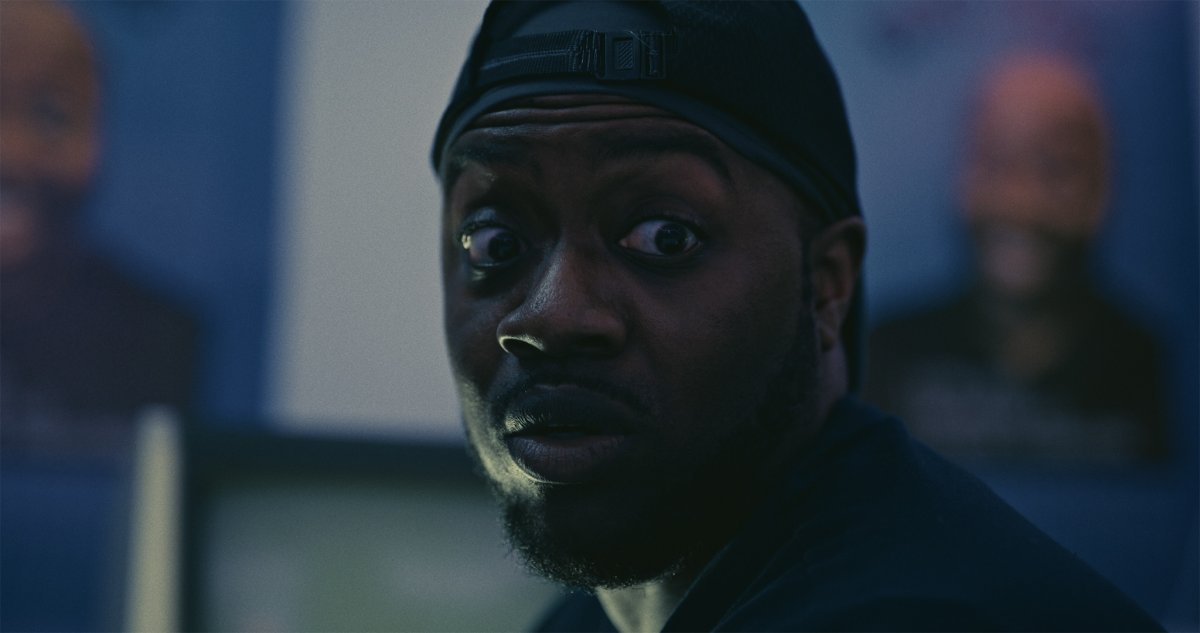 Close-up of a wide-eyed man in a backwards cap staring off-camera in dim blue light