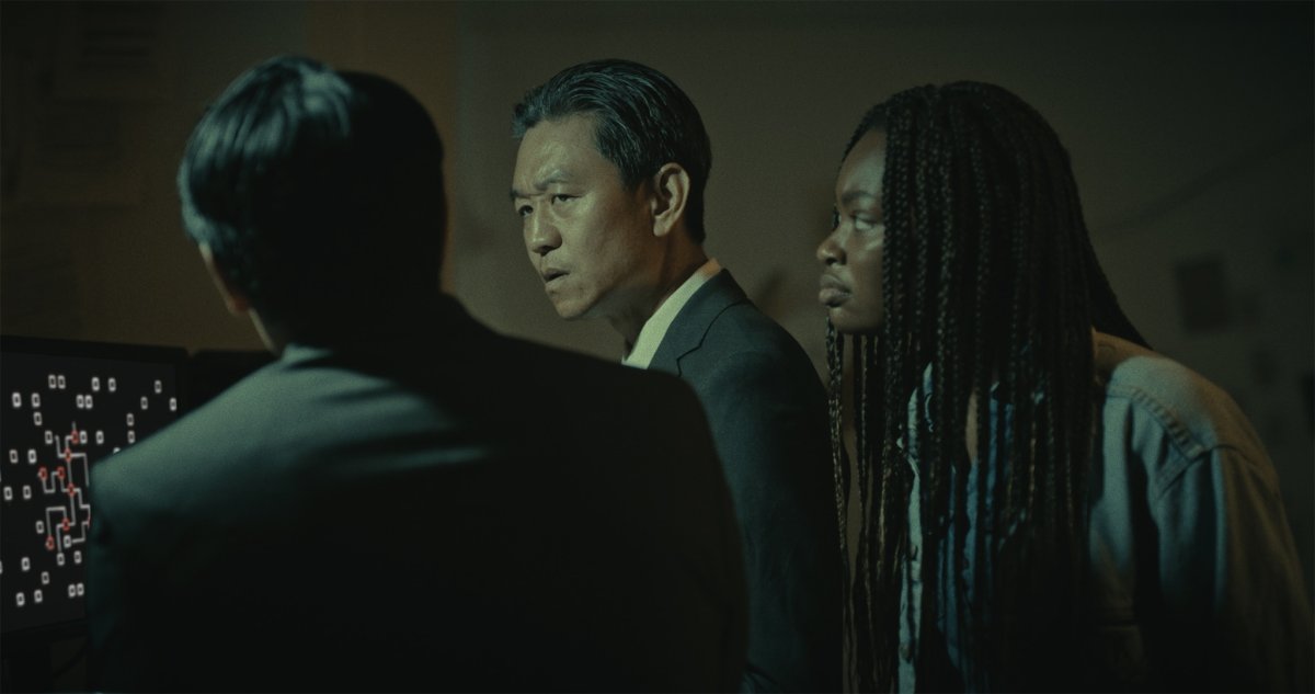 Three people in a dim office look tensely toward something off-frame, a circuit-pattern screen beside them