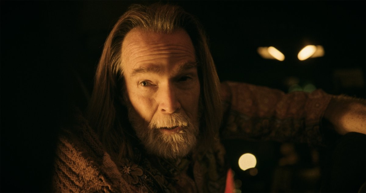 Close-up of a long-haired bearded man lit by warm light, streetlights glowing behind him