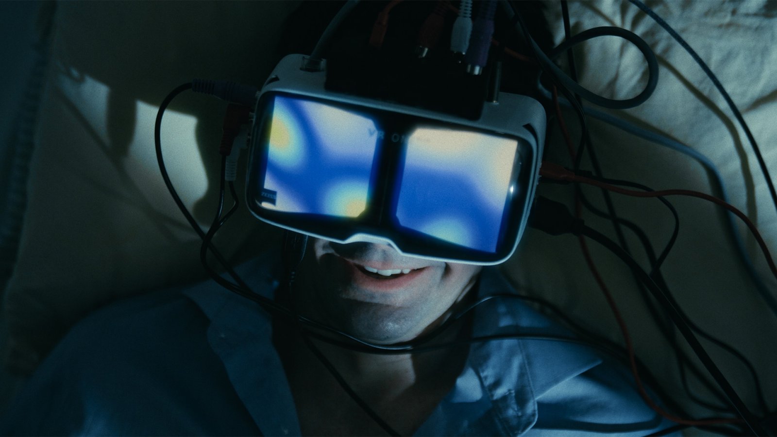 Person lying down wearing a wired VR headset, its glowing blue lenses reflected on their smiling face