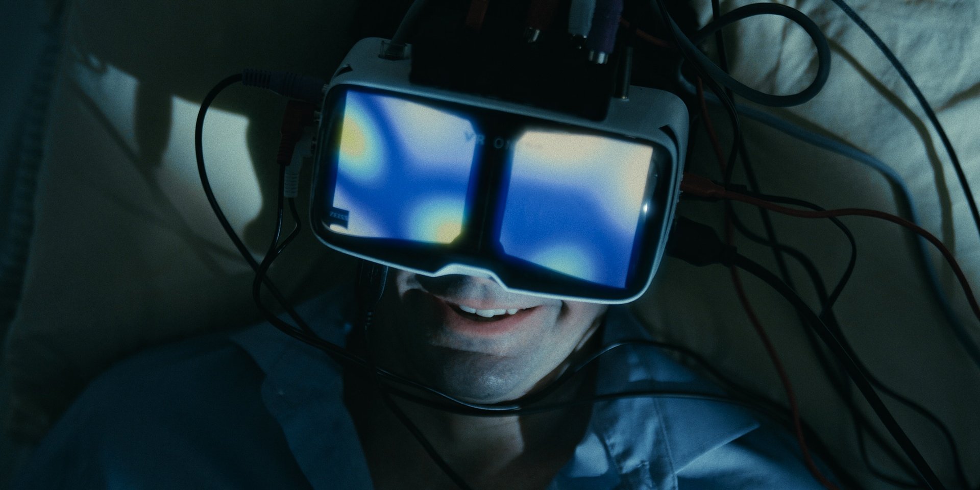 Person lying down wearing a wired VR headset, its glowing blue lenses reflected on their smiling face
