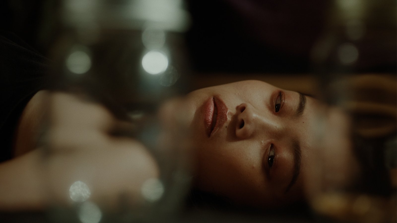 Close-up of a woman’s face reflected upside down in glass, softly lit with bokeh lights in the background