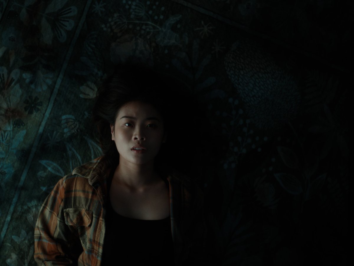 Woman in a plaid jacket lying against floral wallpaper, dim light catching her face