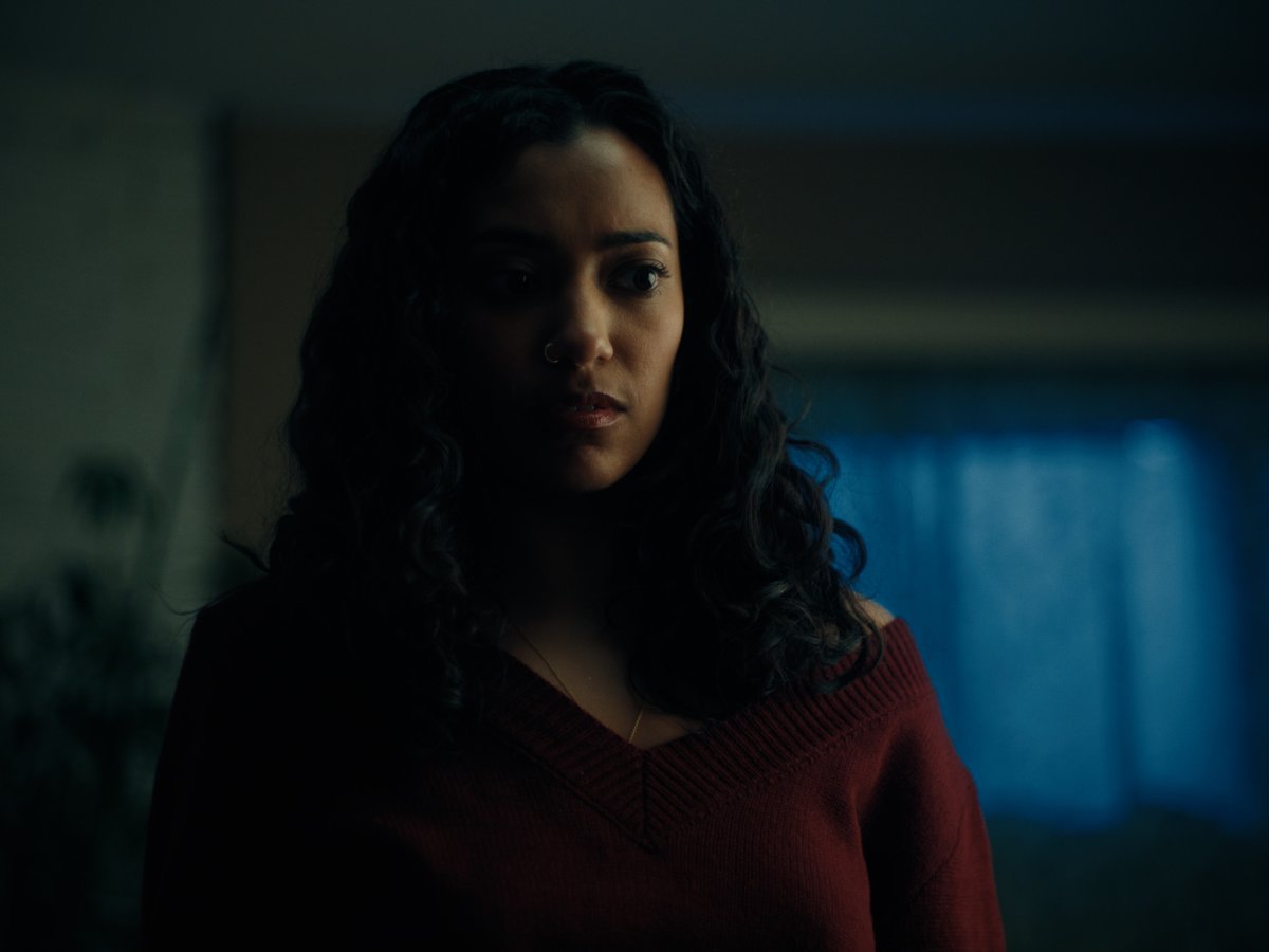 Woman with curly dark hair in a maroon sweater looking off tensely in a dim room
