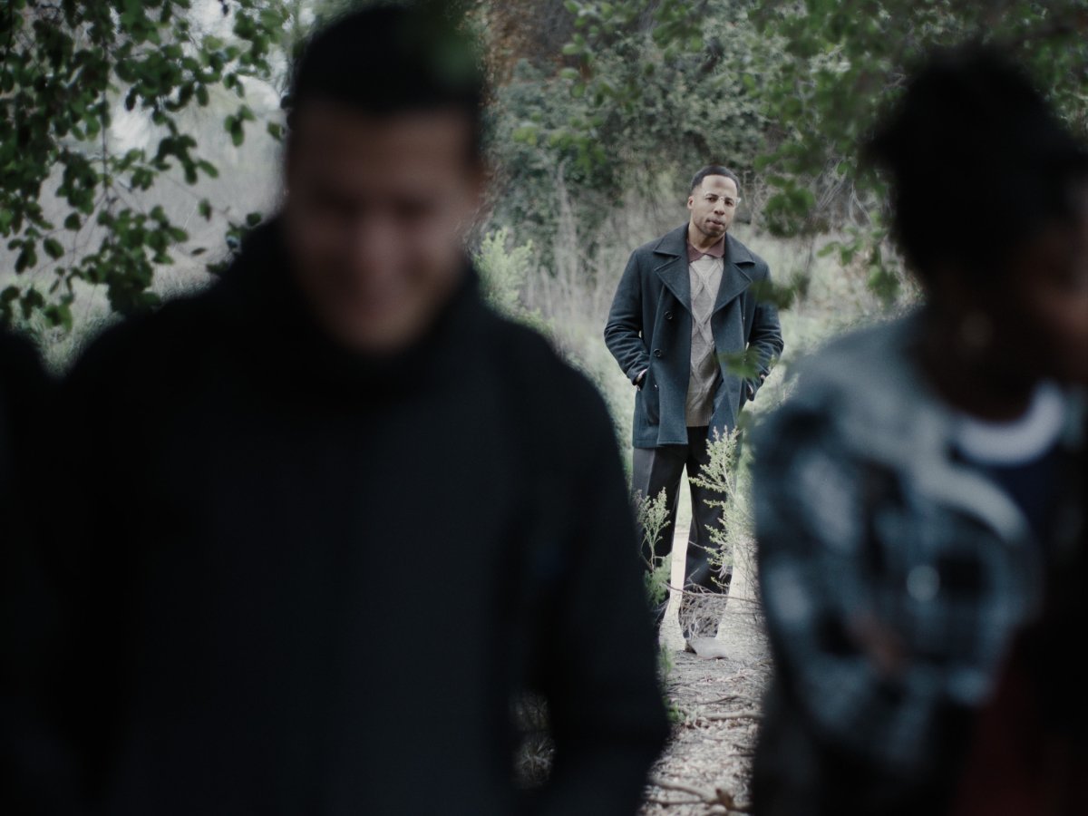 Man in a peacoat standing on a wooded path, two blurred figures in the foreground