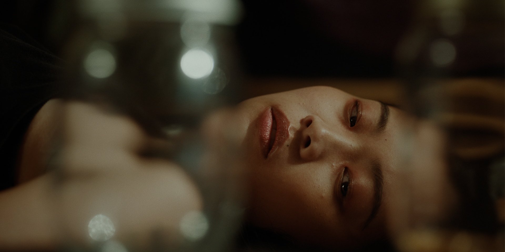 Close-up of a woman’s face reflected upside down in glass, softly lit with bokeh lights in the background