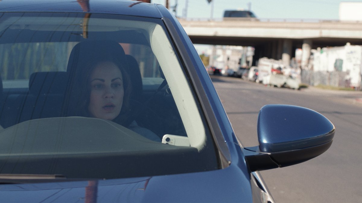 Woman with a startled expression seen through a car windshield, freeway overpass behind her