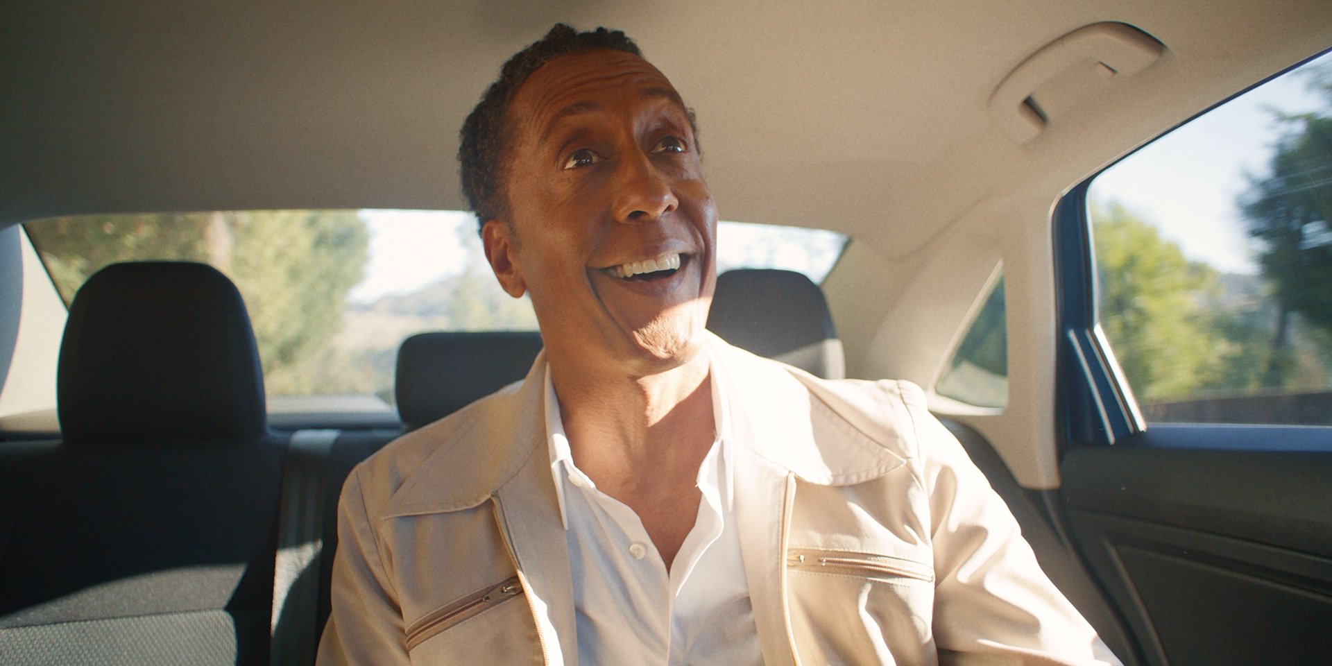 Man in a tan jacket laughing in the back seat of a car, sunlight streaming through the window