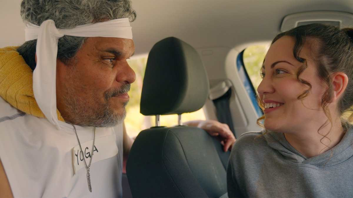 Man in a headband and towel reading "YOGA" smiling at a woman in the back seat