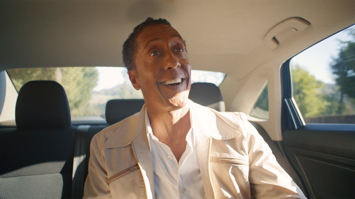 Man in a tan jacket grinning in the back seat of a car, sunlight on his face