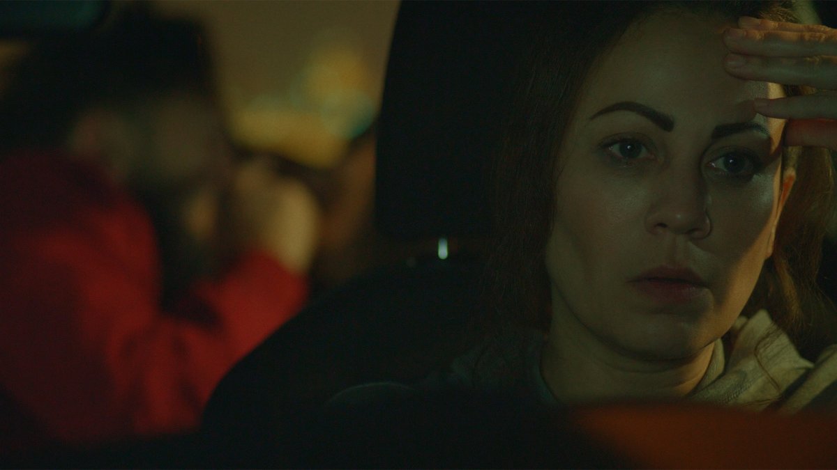 Woman in a car at night shielding her eyes, a figure in a red jacket blurred in the background
