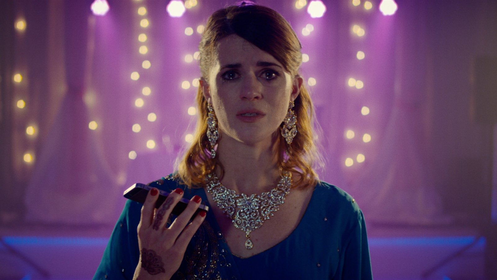 Tearful woman in jeweled earrings and necklace holding a phone, string lights glowing behind her